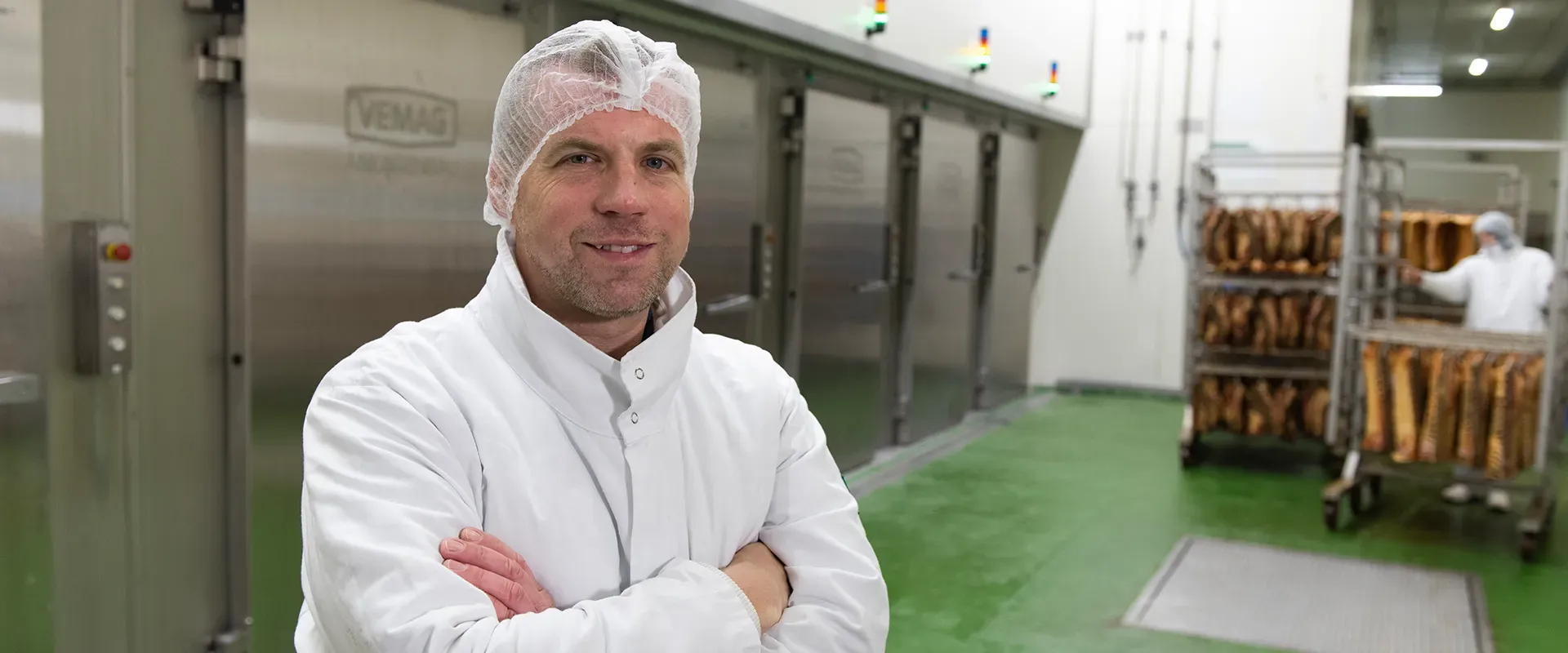 Der Logistik Betriebsleiter von Goldschmaus steht mit verschränkten Armen in einem Durchgang zur Produktion, im Hintergrund sind Wurstwaren zu sehen